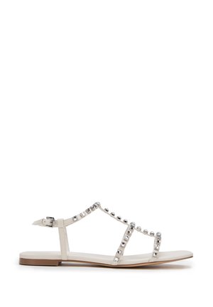Sandal in beige con cinghie decorate con borchie argentate lucide, suola piatta e cinturino alla caviglia per una vestibilità sicura. Design semplice con lati aperti.