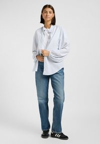 Camicia a righe azzurro chiaro con un fit morbido, maniche lunghe e una t-shirt bianca sotto, abbinata a jeans blu e sneakers nere.