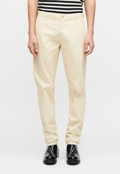 Pantalon en coton beige à coupe droite, avec une braguette zippée et une fermeture par bouton, associé à des bottines noires.