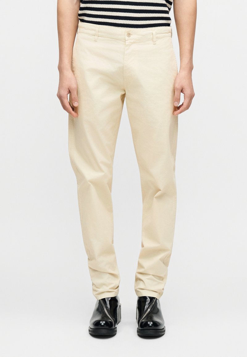 Pantalon en coton beige à coupe droite, avec une braguette zippée et une fermeture par bouton, associé à des bottines noires.