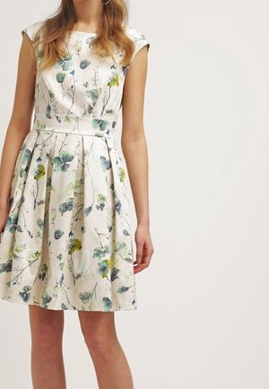 Femme portant une robe blanche sans manches, longueur genoux, avec un imprimé floral vert et bleu et une jupe plissée.