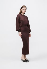 Bordowa sukienka z miękkiej tkaniny o luźnym kroju, ze ściąganym w talii i długimi rękawami. Połączona z czarnymi mule na wysokim obcasie.