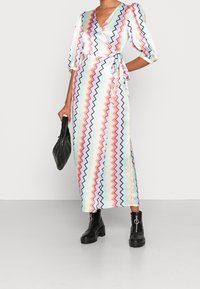 Lång, omlottdress i pastellfärgade zigzag-ränder av rosa, blått och kräm. Har ballongärmar och knytskärp i midjan, matchad med svarta ankelskor.