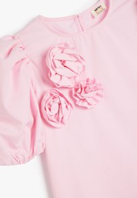 Roze jurk met opgeblazen mouwen, voorzien van drie stoffen rozetten aan de voorkant. Gladde textuur, licht materiaal en ritsluiting aan de achterkant.