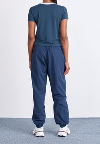 Žena stojící zády, oblečená v tyrkysovém tričku s krátkým rukávem, volných námořnických kalhotách, bílých teniskách a bílých ponožkách na šedém koberci.