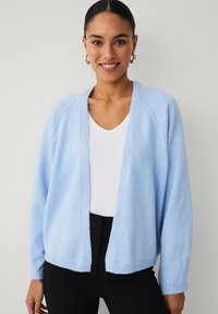 Cardigan en tricot bleu clair à coupe décontractée, à manches longues avec poignets côtelés et ouverture sans bouton. Porté sur un haut blanc et associé à un pantalon noir.