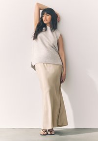 Ljusgrå ärmlös stickad topp med ribbad fåll kombinerad med en luftig beige maxikjol och svarta remsandaler mot en enfärgad bakgrund.