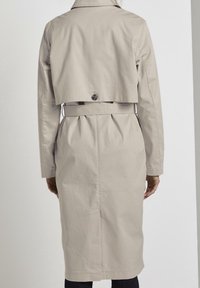 Beżowy trench coat o skróconym fasonie, z zapięciem na guziki z tyłu oraz paskiem w talii. Gładka tekstura, długie rękawy i dopasowany krój.