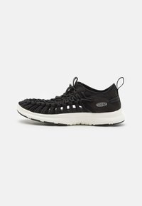 Scarpa sportiva nera con parte superiore intrecciata, suola bianca e linguetta sul tallone. Design leggero con materiale flessibile e traspirante.
