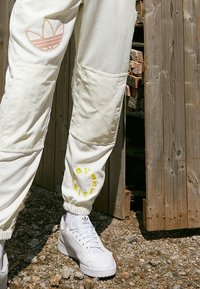 Witte broek met gele tekst en een roze Adidas-logo, gecombineerd met witte Adidas-sneakers, staande op grind naast een houten structuur.