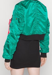 Groene satijnen bomberjack met ingezette mouwen, een zwarte geribbelde tailleband en een rood label. De jas heeft een cropped ontwerp.