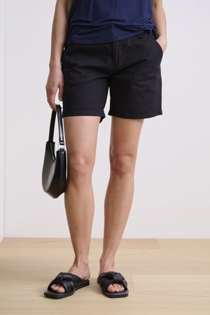 Shorts en coton noirs avec des poignets roulés, associés à des sandales noires à glissière et un sac noir. Le modèle porte un haut bleu marine et se tient sur un sol en bois.