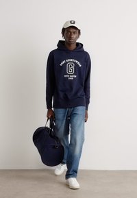 Felpa navy con grafica bianca, jeans blu e sneakers bianche. Tiene una borsa a tracolla blu scuro, indossa un cappellino abbinato. Design semplice.