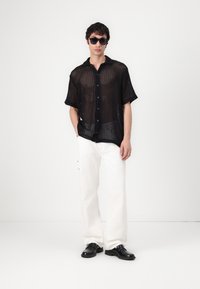 Schwarzes Mesh-Hemd mit kurzen Ärmeln und Knopfverschluss, kombiniert mit weißen weit geschnittenen Hosen und schwarzen Schuhen. Eine Sonnenbrille rundet das Outfit ab.