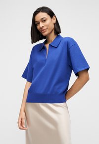 Κοντομάνικη μπλούζα πόλο σε έντονο μπλε πλεκτό ύφασμα, με γιακά και φερμουάρ στη λαιμόκοψη. Μάλλινη υφή με εφαρμοστό κάτω μέρος στη μέση.