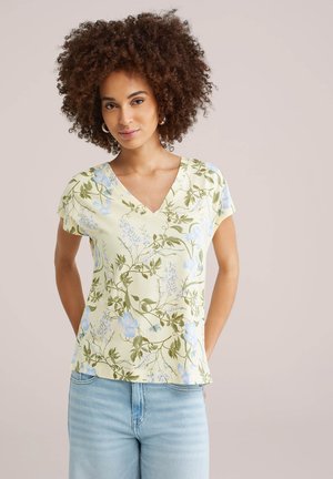 Jonge vrouw met krullend haar, gekleed in een lichtgele blouse met bloemenprint, V-hals en korte mouwen, en een lichtblauwe spijkerbroek, staand met de handen achter de rug.