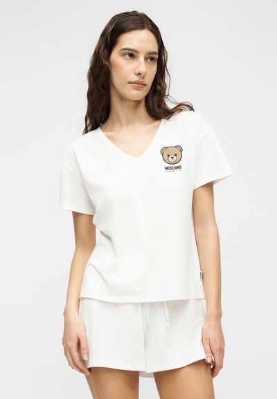 Γυναίκα που φοράει λευκό T-shirt με V-λαιμόκοψη και καφέ λογότυπο αρκούδας, καθώς και λευκά σορτς, στέκεται μπροστά σε απλό φωτεινό φόντο.