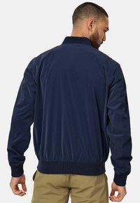 Marineblå bomberjakke med ståkrave, ribbede manchetter og talje; lavet af glat materiale; har et rent, minimalistisk design.