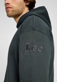 Sudadera verde oscuro con un logotipo texturizado de Lee en la manga izquierda, hecha de una tela suave con un corte relajado y capucha con cordón.