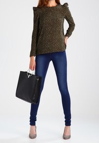 Top verde scuro con motivo floreale bianco, maniche lunghe e spalle arricciate. Abbinato a jeans skinny blu scuro e una borsa nera.