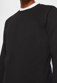 Hombre vestido con un sudadera negra de cuello redondo en capas sobre una camiseta blanca, visible desde el cuello hasta la cintura, contra un fondo blanco liso.