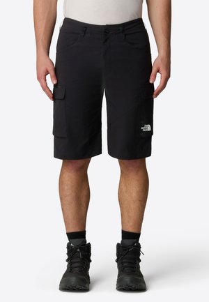 Homme portant un short cargo noir avec poches latérales et des chaussures de randonnée noires, debout devant un fond clair uni.