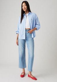 Sieviete stāv ar roku kabatā, uzvilkusi gaismas zila pārāk lielu krekli, baltu crop top, gaismas zila bikses un sarkanas plakanas kurpes uz pelēka fona.