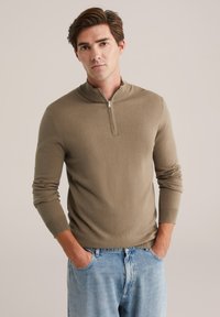 Pulover bej cu fermoar, cu guler înalt, manșete elastice, siluetă ajustată, realizat dintr-un material moale, texturat, purtat peste blugi albastru deschis.