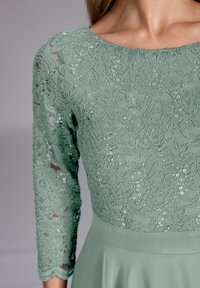 Robe en dentelle verte à manches trois-quarts, ornée de motifs floraux, de sequins et d'une jupe fluide. Le tissu présente une apparence délicate et texturée.