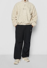 Pullover crema in pile con zip e collo alto, abbinato a pantaloni neri, ampi, con tasche e un dettaglio di logo discreto.