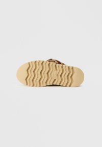 Barna mintás szandál, texturált bézs talppal, amelyen hullámos tapadású minta található, és puha, szőrszerű felső anyag jellemzi.