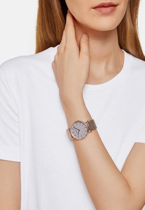 Mesh-båndur med en sølvskive, minimalistiske time markører og en rosaguld ramme; båret på et håndled mod en simpel hvid t-shirt.