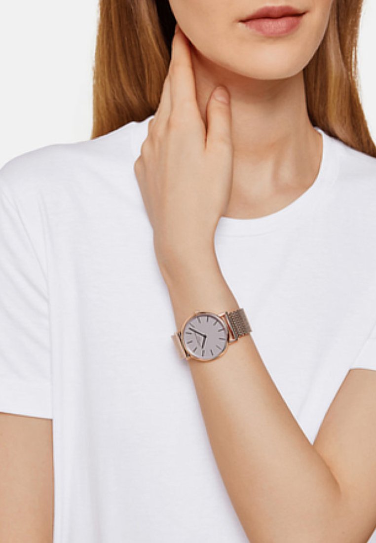 Armbanduhr mit Mesh-Band, silbernem Ziffernblatt, minimalistischen Stundenmarkierungen und einem roségoldenen Gehäuse; am Handgelenk über einem schlichten weißen T-Shirt getragen.