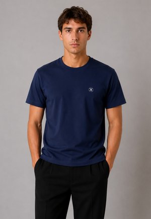 Jeune homme debout les mains dans les poches, portant un t-shirt bleu marine avec un petit logo cercle blanc et croix sur la poitrine et un pantalon noir.