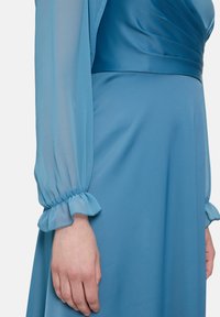 Robe bleu clair avec de longues manches transparentes à volants, un corsage ajusté et une jupe en tissu fluide.