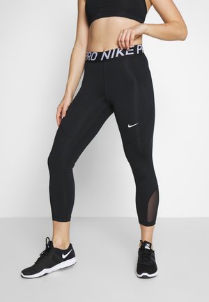 Frau trägt schwarze Nike Pro Leggings und schwarze Nike Sneakers, steht mit einer Hand am Bund und die Beine leicht auseinander.