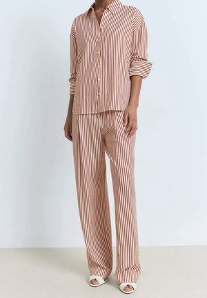 Kobieta ubrana w luźną koszulę na guziki w pionowe beżowo-czerwone paski oraz pasujące do niej szerokie spodnie z białymi sandałami na odkryte palce.