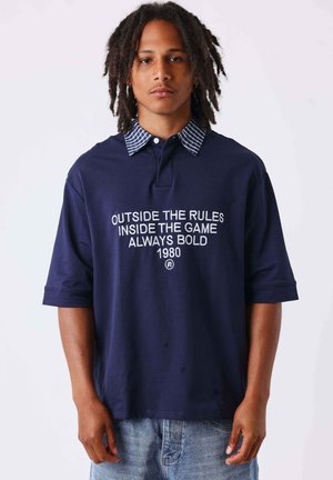 Jeune homme avec des dreadlocks portant une chemise bleu marine avec un col rayé et le texte "Outside the rules inside the game always bold 1980" se tenant devant un fond blanc.