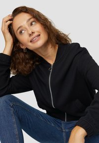 Schwarze Zip-Hoodie aus strukturiertem Stoff, mit elastischen Bündchen und einem glatten Reißverschluss. Kombiniert mit dunkelblauen Jeans.