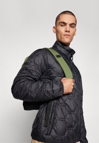 Sort, quiltet pufferjakke med hexagonal mønster, høj krave og dobbelte lynlåslommer. Grøn skulderrem fra en sort taske.