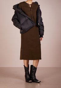 Abito a maglia verde oliva con scollatura rotonda e dettagli lacciati, sovrapposto a un giubbotto imbottito nero. Stivali cowboy neri con punta e accenti argentati.