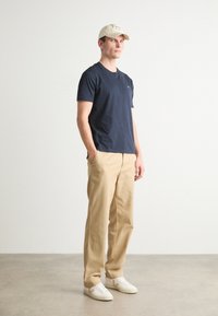 Mørkeblå kortærmet t-shirt, lysebrune afslappede bukser og hvide sneakers. Kasketten er beige med tekst. Modellen står foran en ensfarvet baggrund.