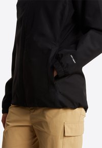 Άτομο που φοράει μαύρο μπουφάν DRYVENT με φερμουάρ σε πλαϊνή τσέπη και μπεζ παντελόνι καμπαρντίνας, χέρι τοποθετημένο στην τσέπη του μπουφάν.