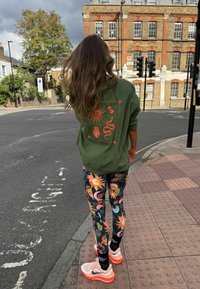 Zelená oversize mikina s oranžovými grafickými motívmi, skombinovaná s čiernymi legínami s kvetinovým vzorom a bielo-ružovými športovými topánkami. Mestská pouličná scéna.