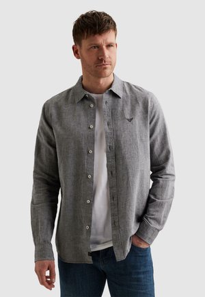 Homme portant une chemise grise boutonnée sur un t-shirt blanc, une main dans la poche du jean, regardant sur le côté devant un fond uni.