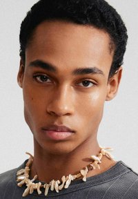 Beige kralenketting gemaakt van koraalachtige vormen, liggend op een grijze, geribde crewneck trui. Close-up van gezichtskenmerken en huidstructuur.