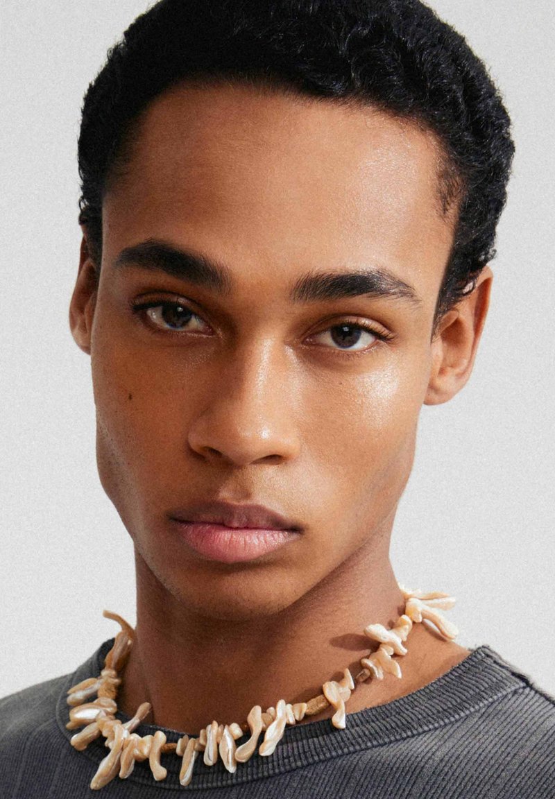Beige kralenketting gemaakt van koraalachtige vormen, liggend op een grijze, geribde crewneck trui. Close-up van gezichtskenmerken en huidstructuur.