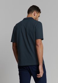 Man in een donkerblauwe korte mouwen polo en marineblauwe broek, met zijn rug naar de camera gericht tegen een effen lichtgrijze achtergrond.