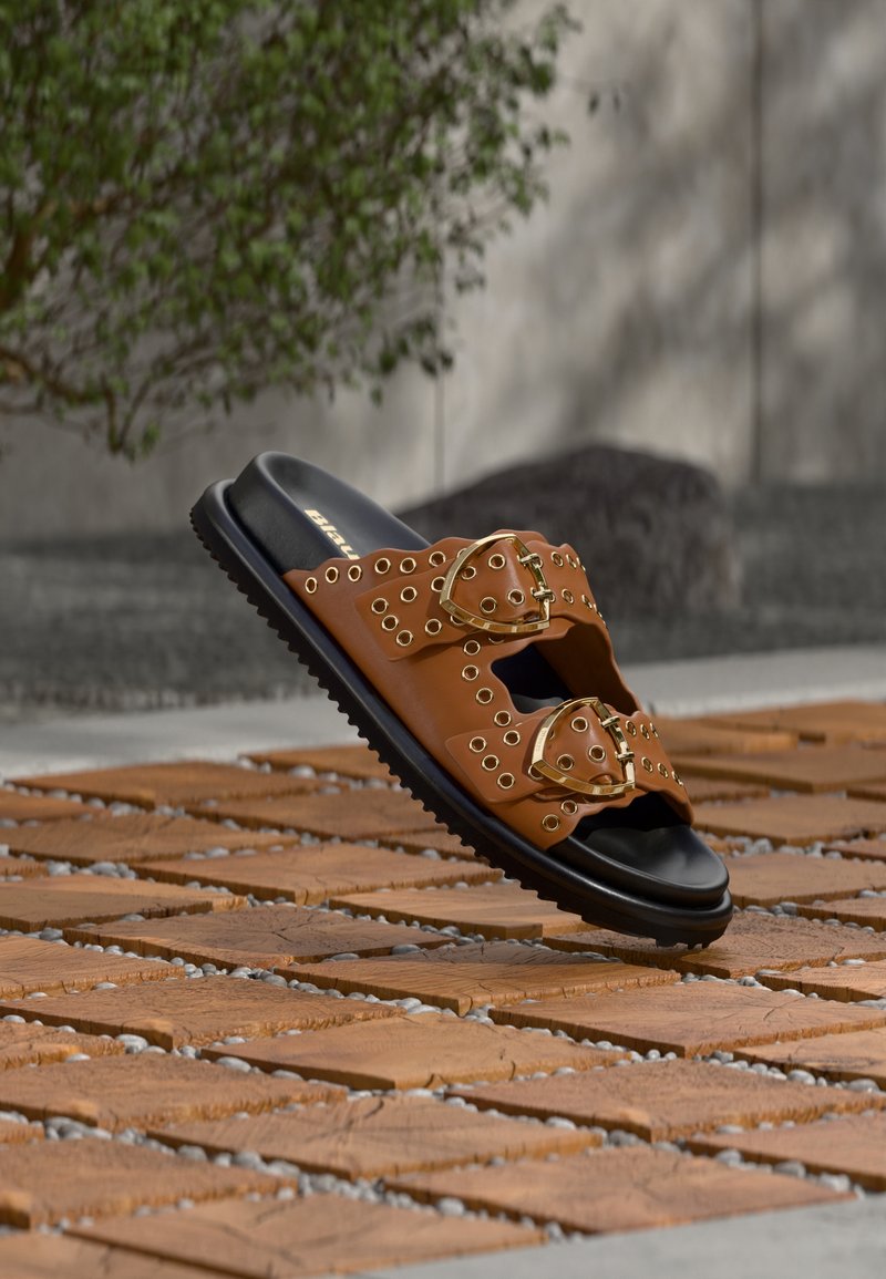 Καφέ δερμάτινο σανδάλι slide με δύο μεγάλες αγκράφες σε χρυσό τόνο, λεπτομέρειες με χρυσές οπές και μαύρη υφασμάτινη λάστιχη σόλα.