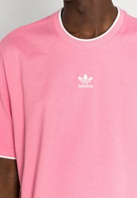 Persona di carnagione scura che indossa una t-shirt Adidas rosa con bordi bianchi e un piccolo logo Adidas bianco sul petto.